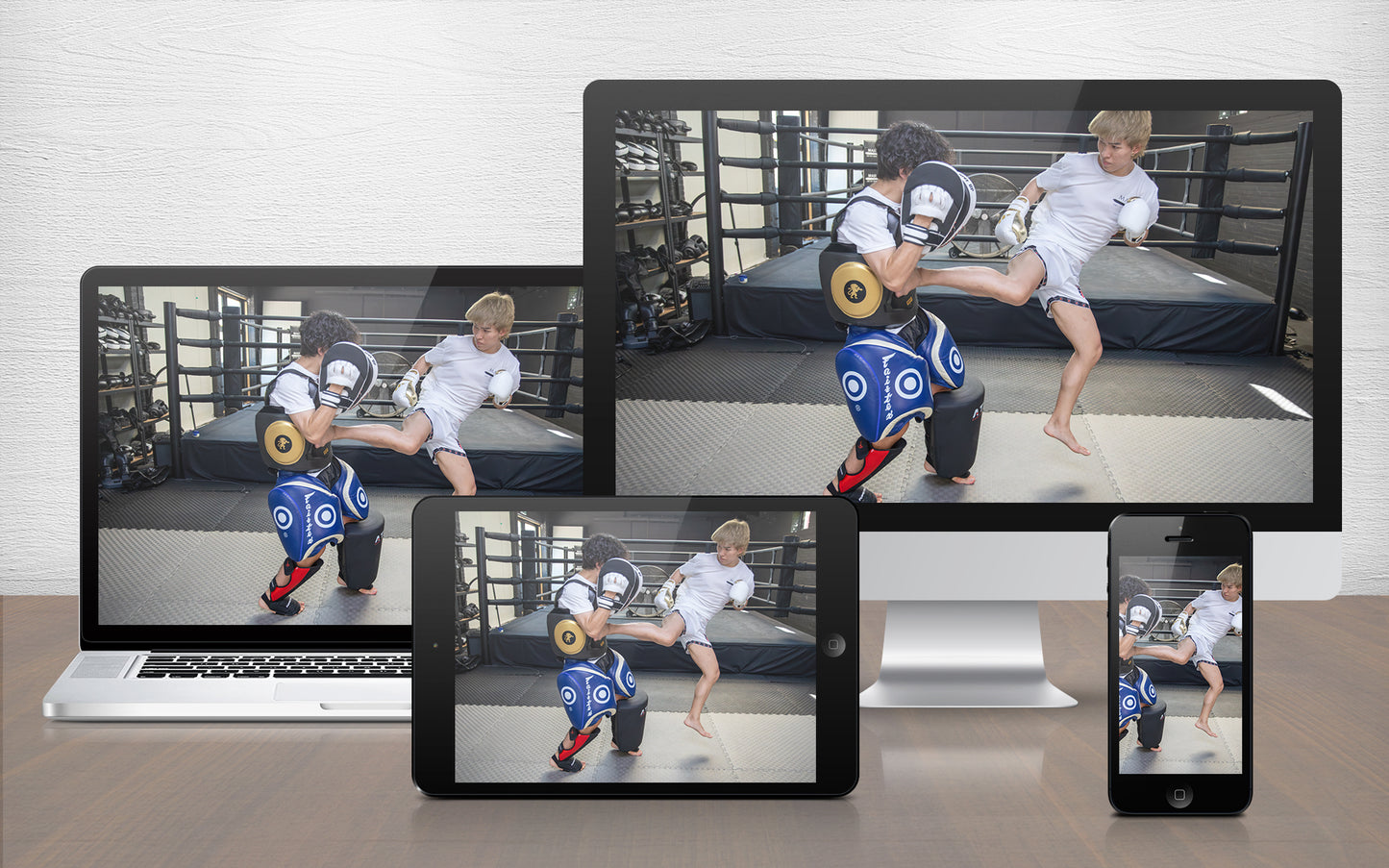 A digital martial arts training program displayed across multiple devices including a desktop monitor, laptop, tablet, and smartphone, showing a video of a fighter practising kicks in front of a boxing ring.