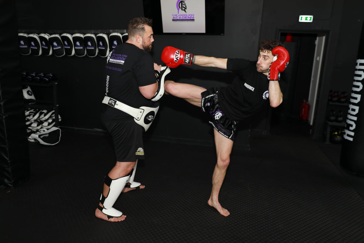 Two men in a martial arts training gym, one wearing a black Muay Thai gi and kicking the other man's head with a white and black Muay Thai pad. The gym has black walls, a rack of Muay Thai shoes on the left, and a black punching bag on the right.