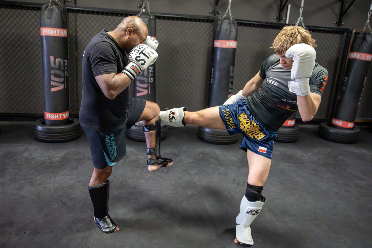Two individuals training in a boxing ring with punching bags in the background.
