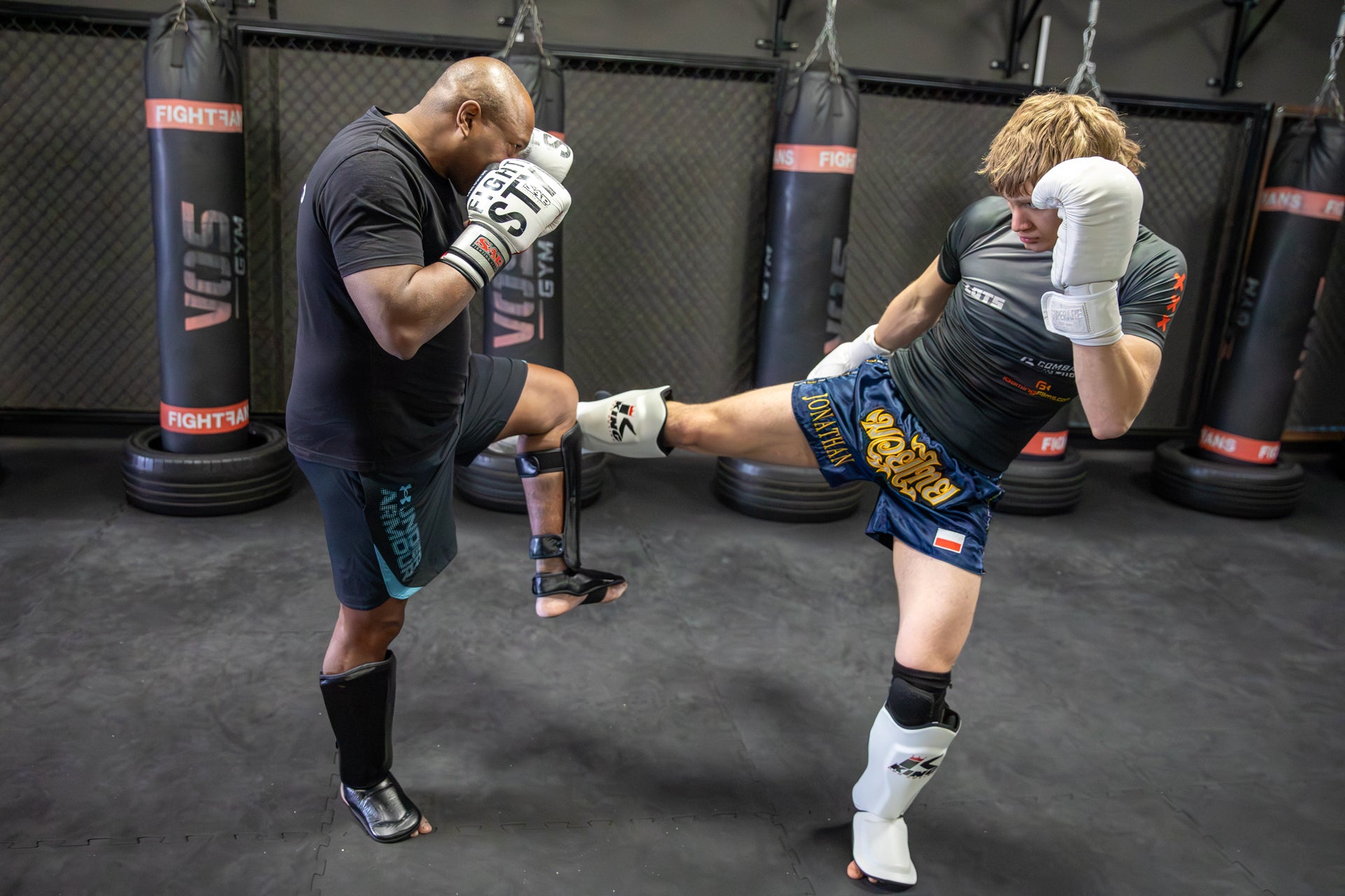Two individuals training in a boxing ring with punching bags in the background.