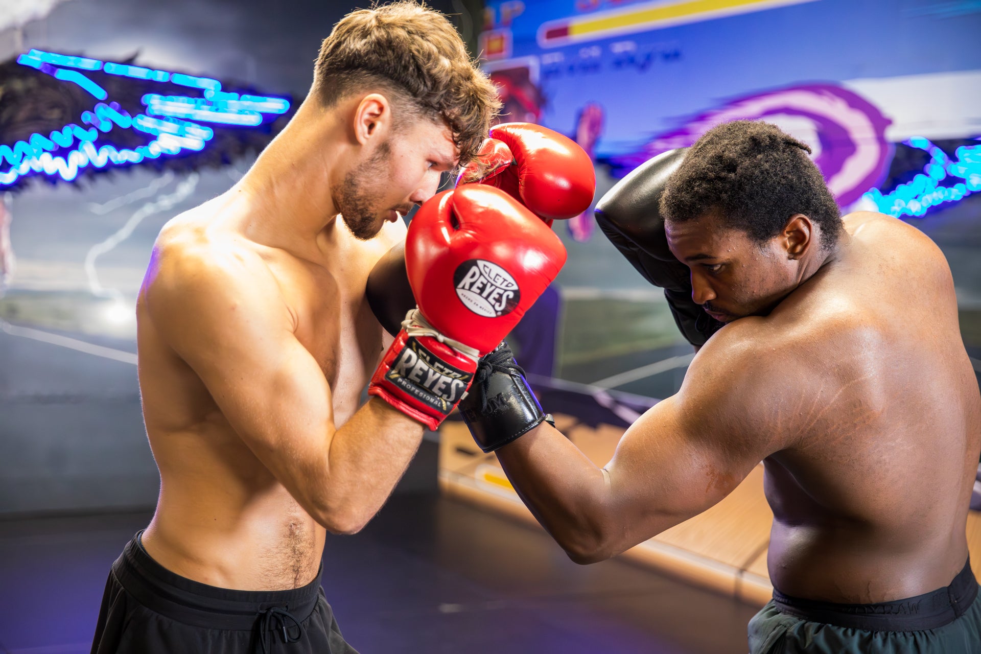 Two fighters training, one wearing red Everlast boxing gloves and one wearing black gloves punching the other man's head. The background shows blue neon lights.