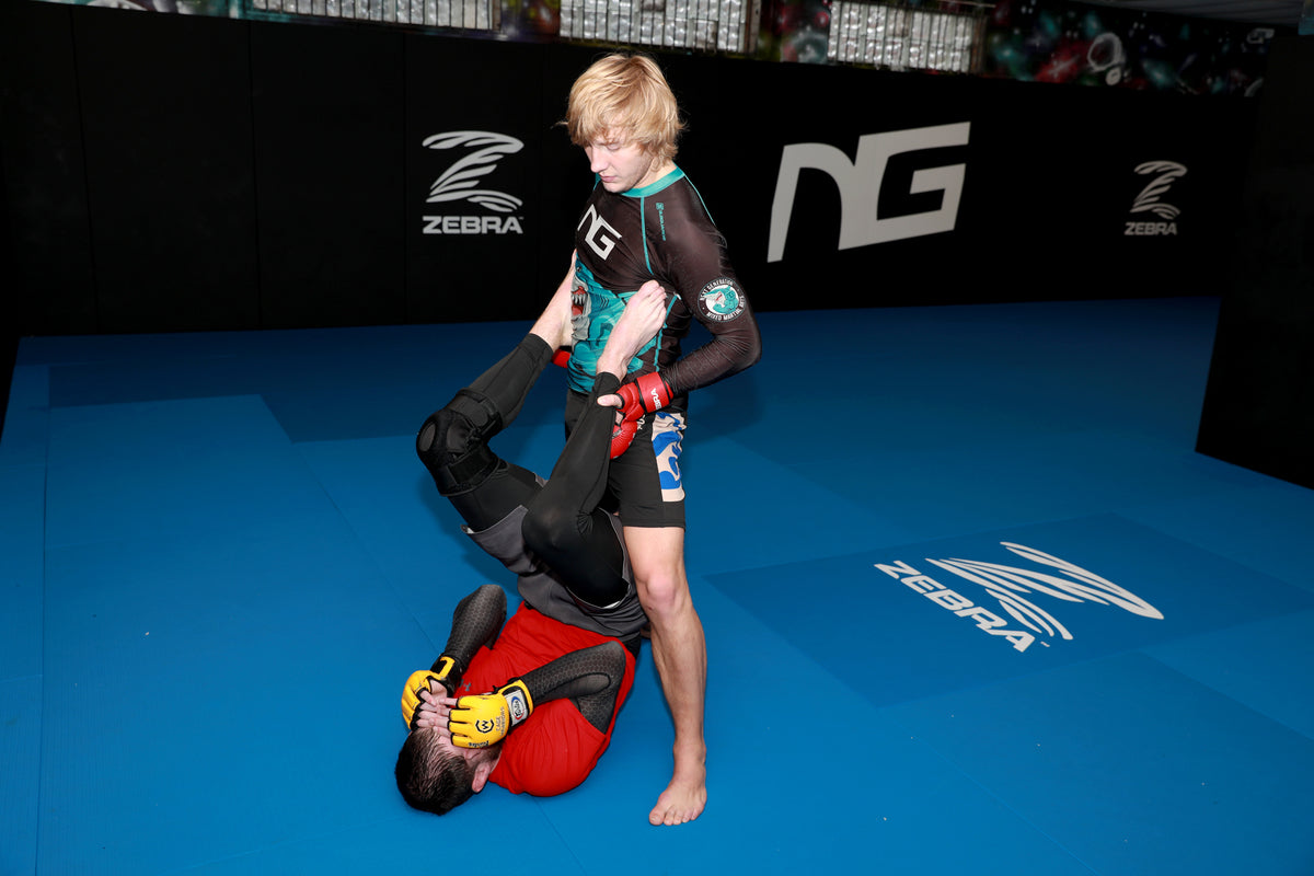 Two martial artists in a blue MMA matted area, with one standing fighter in a black and teal rash guard looking to pass the guard of a fighter in a red rash guard on the mat. Zebra and NG logos are visible on the background wall.